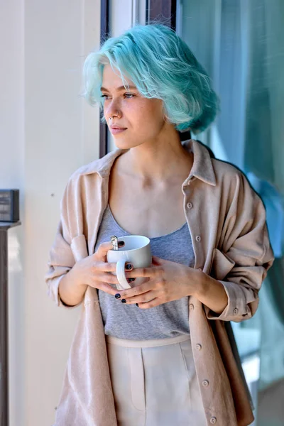 Femme caucasienne calme avec des cheveux bleus inhabituels sur près de fenêtre avec tasse — Photo