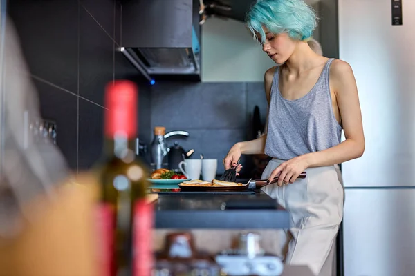 Attractive young woman with modern green hairstyle is cooking breakfast, side view — Stock Fotó