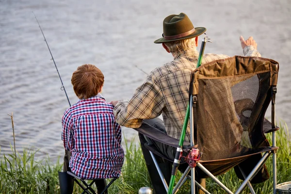 Rear view on mature man with grandson sitting near river with fishing rods in hands — Stock Photo, Image