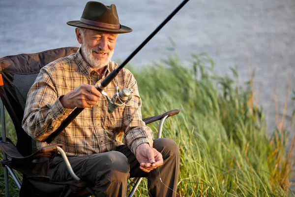 Elderly happy man fishing outside in evening on lake in summer sitting on chair
