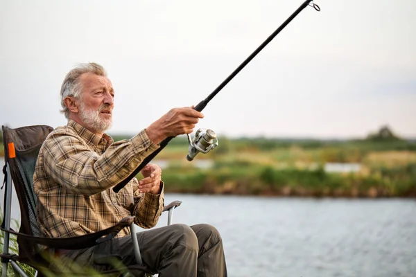 Elderly man fishing outside in evening on lake in summer sitting on chair — Fotografia de Stock