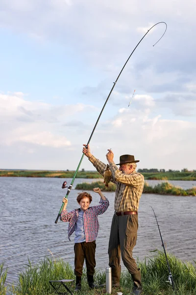 Cheerful little boy holding fishing rod and fish posing next to grandfather in the evening — Fotografia de Stock