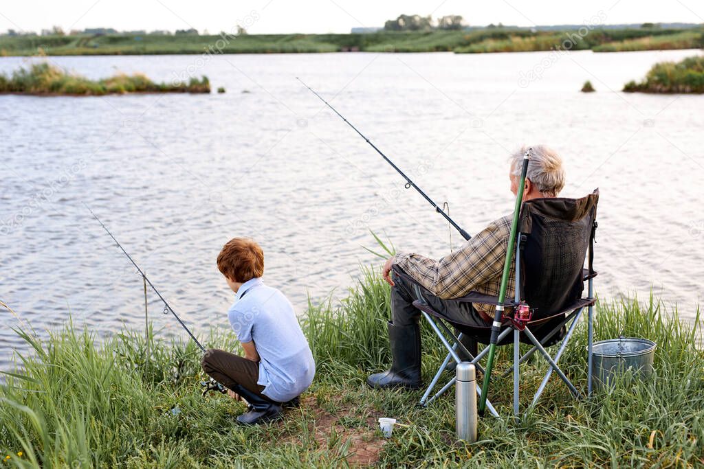 Mature man and grandson fishing on river or lake, having thermos with tea