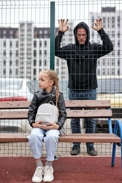 Young maniac guy watches through the fence at little girl sitting on playground alone — Fotografia de Stock