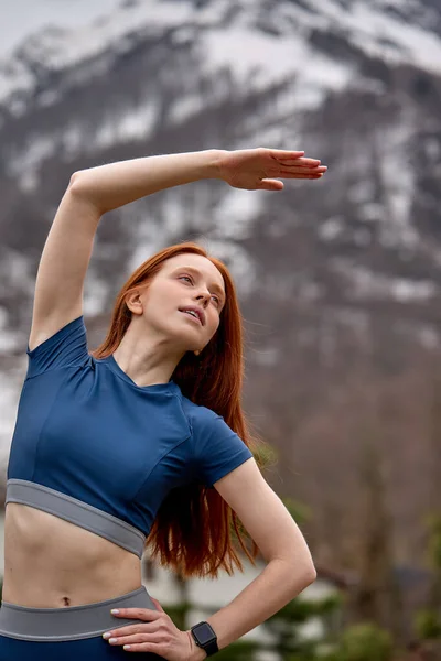 Fitness female in sportive outfit stretching body before workout, raising hands up