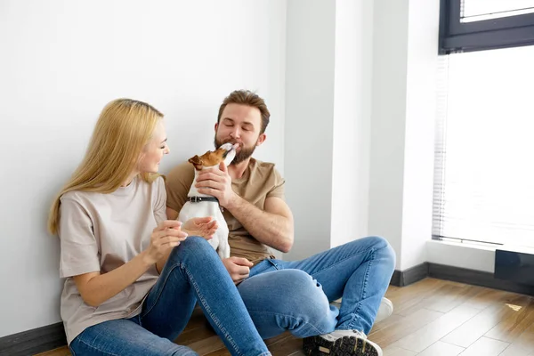 Joven feliz pareja se está moviendo en nueva casa, sentado en el suelo con poco cachorro — Foto de Stock