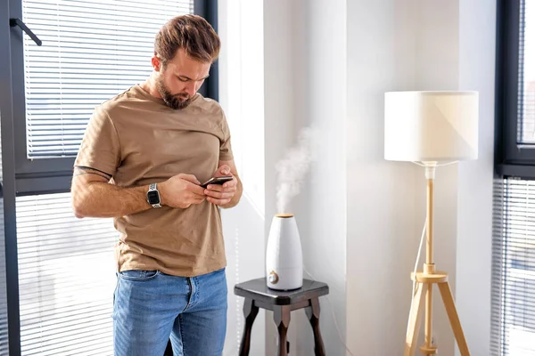 Intelligenter Luftbefeuchter emittiert Wasserdampf auf dem Tisch, während der Mensch das Smartphone nutzt — Stockfoto