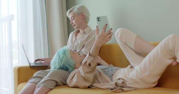 Akıllı telefon kullanan, dizüstü bilgisayarlı, kanepede oturan beyaz, güzel, lezbiyen bir çift. — Stok video