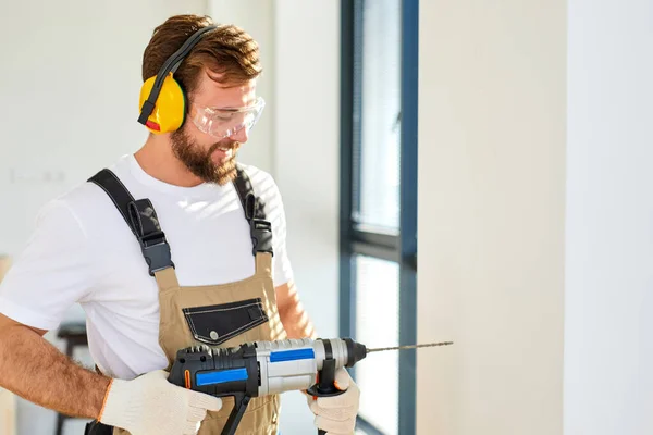 Bouwvakker maakt gat in witte muur met elektrische boor, zijaanzicht — Stockfoto