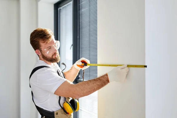 Handyman reparador construtor em macacões e luvas mede parede branca com ferramenta de fita — Fotografia de Stock