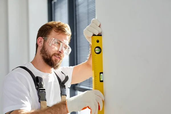 Vista lateral del hombre joven utilizando el nivel en la pared blanca en casa, reparador se concentra en el trabajo —  Fotos de Stock