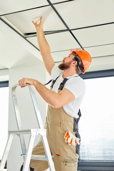 Jovem construtor caucasiano na escada rolante reparação teto, em capacete e macacão — Fotografia de Stock