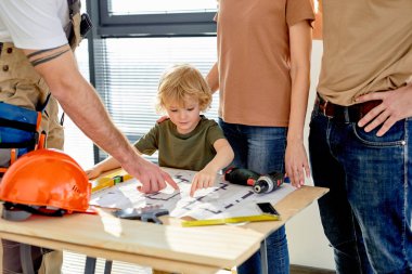 Çocuk tamir detaylarını öğreniyor, ebeveynler inşaatçıyla konuşurken planlara bakıyor.