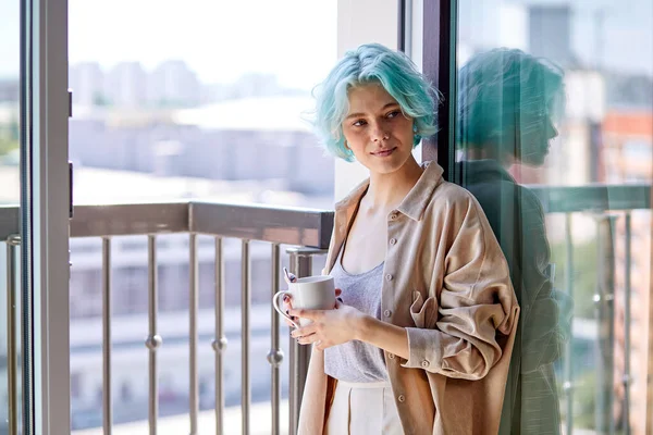 Jeune femme caucasienne avec des cheveux bleus colorés inhabituels sur près de fenêtre avec tasse — Photo
