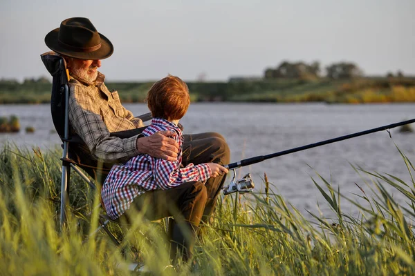 Side View On Happy Mature Man With Grandson Holding Rod While Sitting On River Bank — Stock Photo, Image