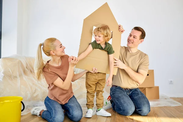 Joyous family and kid boy playing in new home with cardboard boxes on floor. Portrait