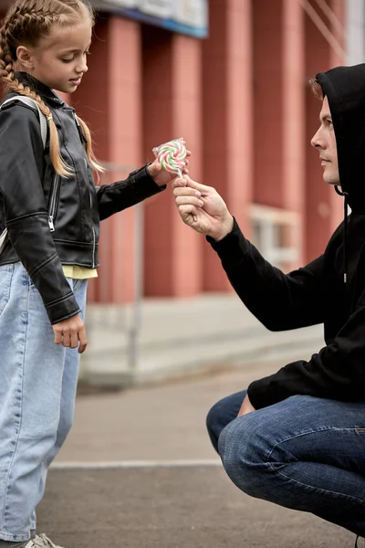 Young man kidnapper with evil face offering lollipop to child girl on city street