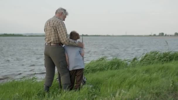 Старший мужчина с внуком вместе пришли ловить рыбу вечером на закате в летний день — стоковое видео