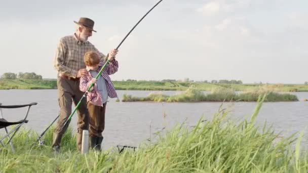Łowienie szczupaków, okoni, karpi. Wykwalifikowany starszy rybak i chłopiec z wędką, kręcący się kołowrotek na brzegu rzeki — Wideo stockowe