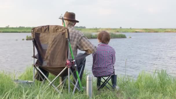Granddad and Grandson During Fishing On River. Sit together in field with fishing rod — Stock Video