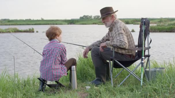 Abuelo y nieto durante la pesca en el lago. Sentarse juntos en el campo con caña de pescar — Vídeo de stock