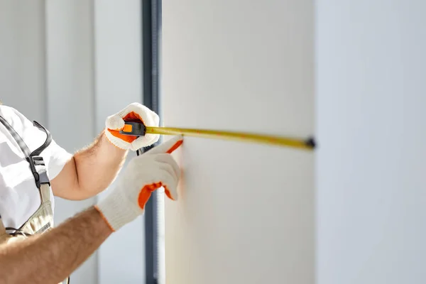 Homem reparador construtor em macacões e luvas mede parede branca com ferramenta de fita — Fotografia de Stock