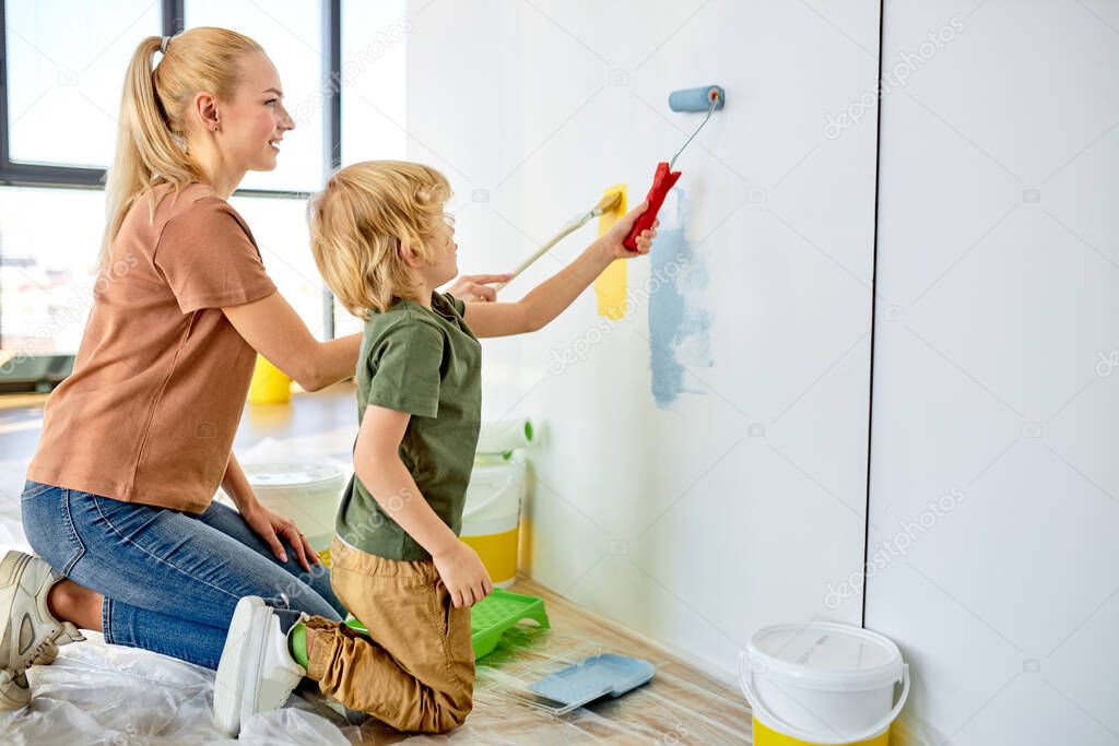 happy mother teaching little adorable kid boy how to paint wall using paintbrush roller