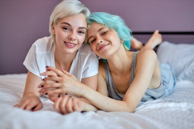 Gülümseyen lezbiyen LGBT çifti yatak odasında sıcacık sarılıyor ve gülüyor.