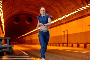 Kızıl saçlı genç bir bayanın portresi. İleriye doğru koşuyor, egzersiz yapıyor ve yol boyunca tek başına antrenman yapıyor.