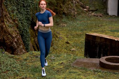 Mavi spor elbiseli aktif kızıl saçlı kadın doğada koşmaktan hoşlanıyor.