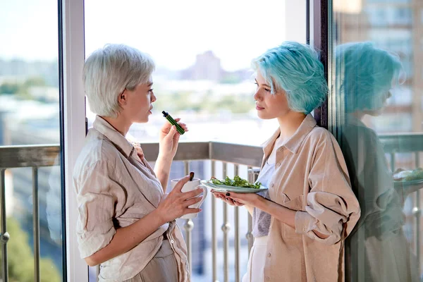 Atractiva pareja de lesbianas de pie en el balcón hablando, fumando y desayunando —  Fotos de Stock