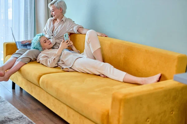 Loving young lesbian couple having free time at home on sofa, using smartphone and laptop — Stock Photo, Image