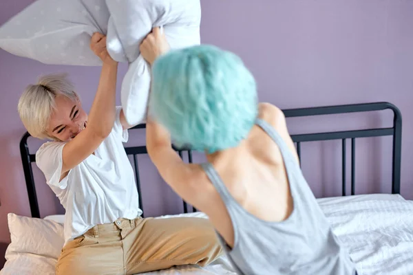 Dost canlısı iki beyaz kadın beyaz yatakta oturmuş yastıklarla oynuyorlar. — Stok fotoğraf