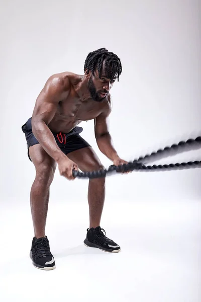 Entrenamiento masculino negro sin camisa enfocado con la cuerda de batalla en el estudio sobre fondo blanco —  Fotos de Stock