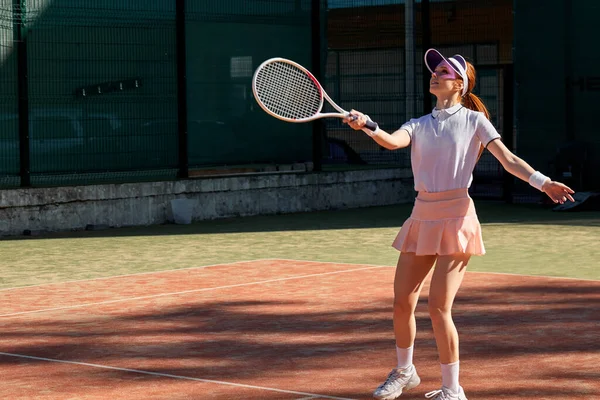 Beyaz tenis üniformalı genç bir bayan tenis oynuyor, raketle topa vuruyor.