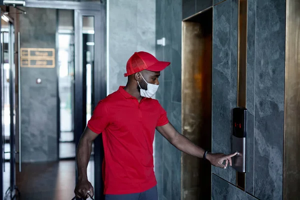 Handsome delivery man in red uniform pushing elevator button in entrance of house — Stock Photo, Image