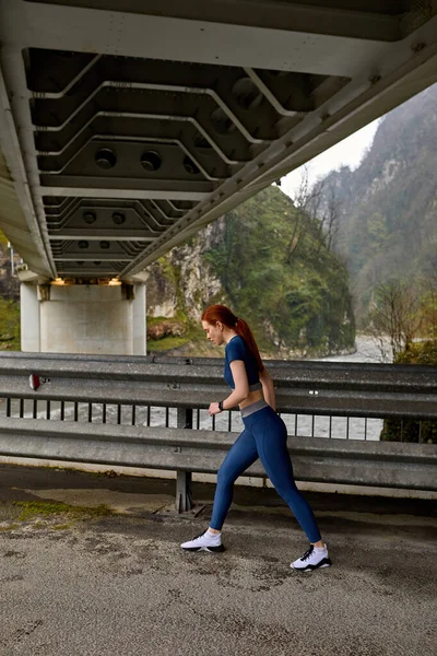 Πλαϊνή άποψη για λεπτή κοκκινομάλλα γυναίκα τρέχει σε ωραίο τοπίο της φύσης, στη γέφυρα — Φωτογραφία Αρχείου