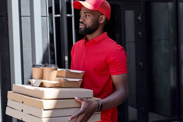 Jovem homem de entrega africano barbudo em uniforme vermelho com caixas de pizza, lancheira e café — Fotografia de Stock