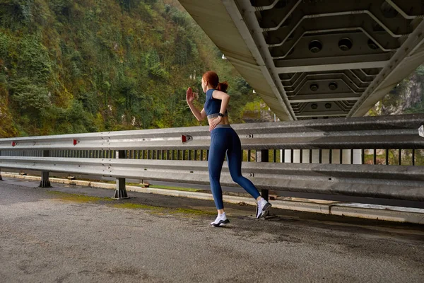 Zijaanzicht op actieve roodharige vrouw rennend in mooi natuurlandschap, op brug — Stockfoto