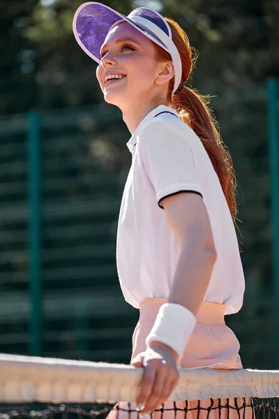 Side View Portrait Of Excited Female In Tennis Female Uniform Standing By Net