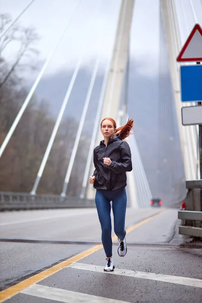 都会のスポーツで健康な若い女性が寒い春の朝に街をジョギングし — ストック写真