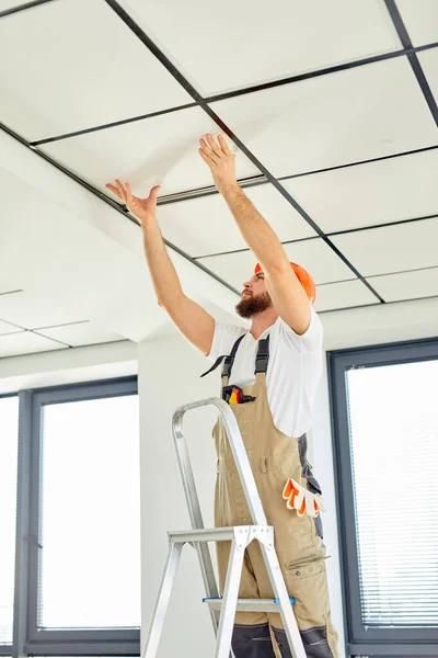 Professionele bouwer op trapladder die plafond repareert, in helm en overall — Stockfoto