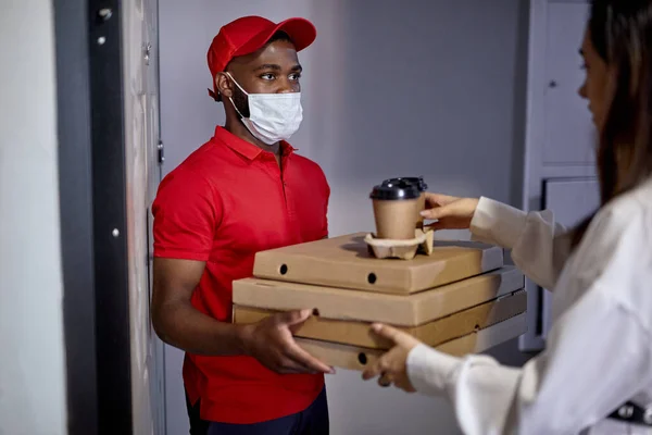Mensageiro masculino africano em máscara médica trazendo pizza e café para a jovem. — Fotografia de Stock