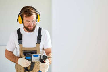 Kulaklıklı inşaat işçisi, beyaz duvarda elektrikli matkapla delik açıyor.