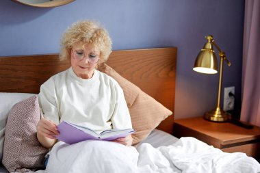 Kitap rahatlamak için iyidir. Evde yaşlı beyaz bir kadın oturuyor ve kitap okuyor.