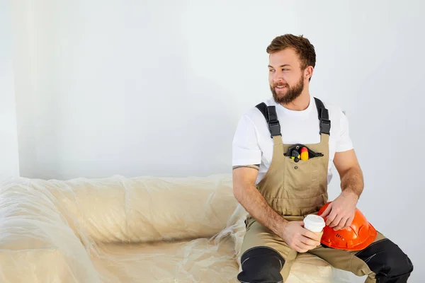 Handsome young caucasian builder is drinking coffee on break time, sits smiling. — Stock Photo, Image