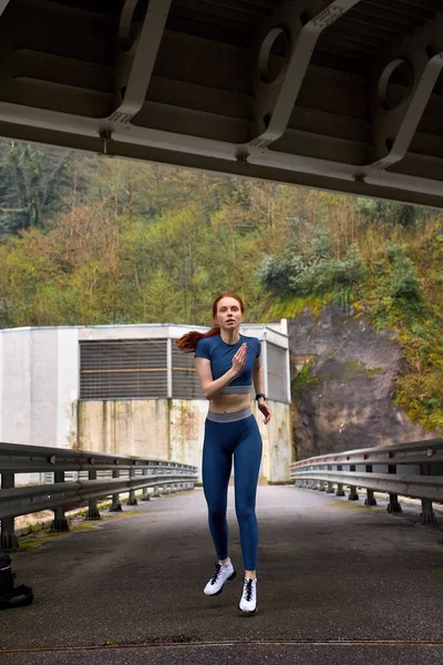 Foto de Portrait of confident female runner jogger training outdoors on  bridge alone, doing exercises, in sportive outfit, in the morning, cold  day. workout, sport, jogging, healthy lifestyle concept do Stock
