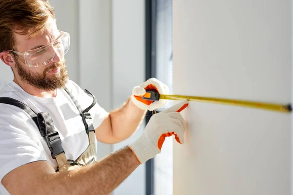 Handyman reparador construtor em macacões e luvas mede parede branca com ferramenta de fita — Fotografia de Stock