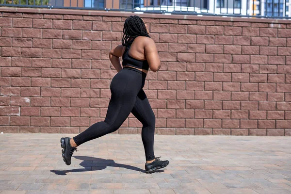Overweight young african woman jogging outdoors. Fat beautiful female in black tracksuit — Stock Photo, Image