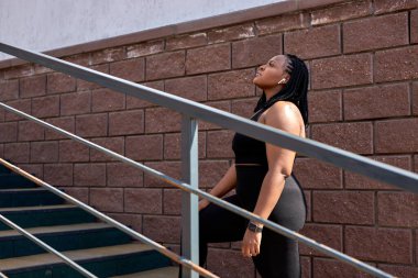 Spor kıyafetleri içindeki genç Afrikalı kadın spor sonrası merdivenlerde dikiliyor.